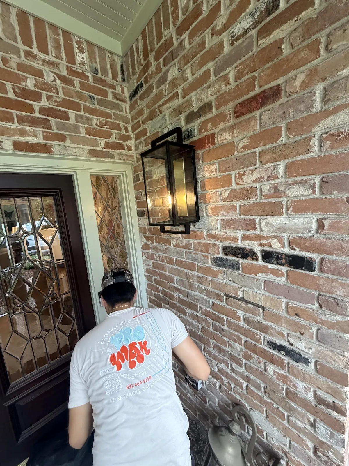 Pro Max Electrical technician in a company shirt installing a lantern light beside a front door on a brick house
